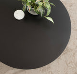 Black round coffee table surface with potted leafy green plant and small white marble container, on beige textured floor.