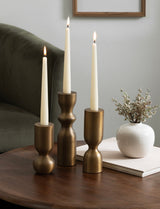Three bronze-toned metal taper candle holders of varying heights with lit white candles, arranged on a wooden table next to a white round vase with dried flowers.