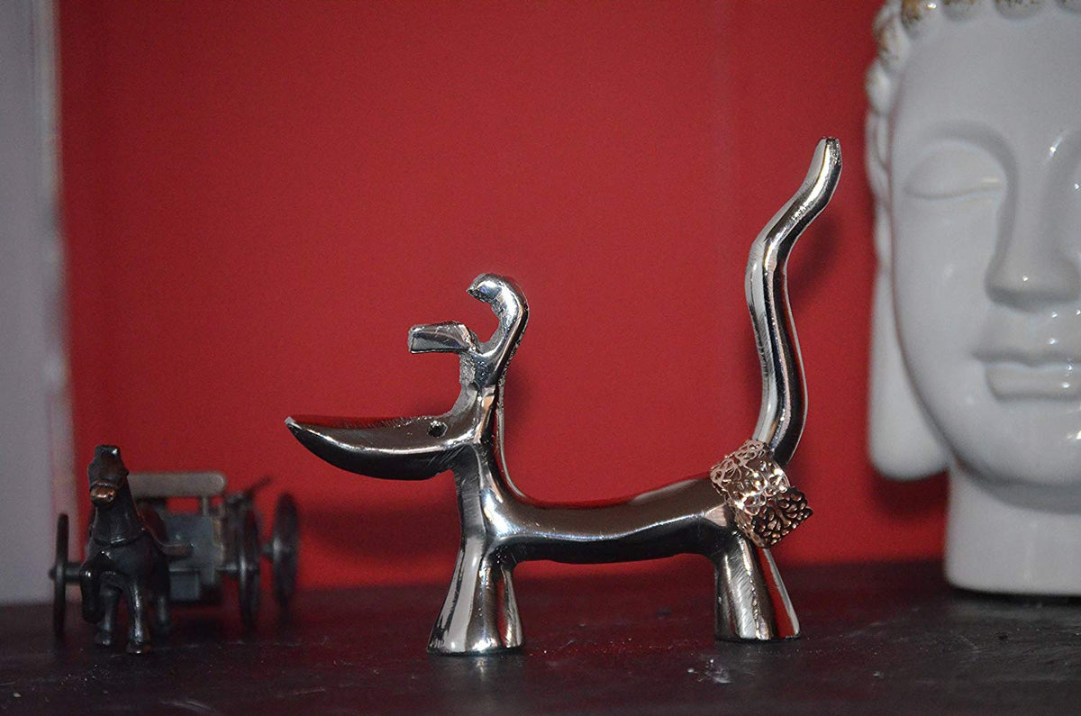 Silver-finished dog-shaped ring holder with two rings on its tail, positioned on a dark surface against a red background with decorative objects nearby.