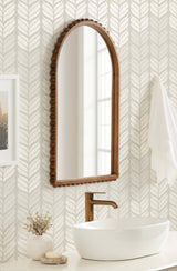 Tall arched mirror with a carved wooden bead frame above a white oval vessel sink and bronze faucet, set against light wallpaper with a leafy pattern.
