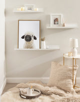 Framed canvas print of a black-nosed sheep with white wool displayed on a wall shelf amid neutral-toned decor including candles, a knitted pillow, and a softly lit lamp.