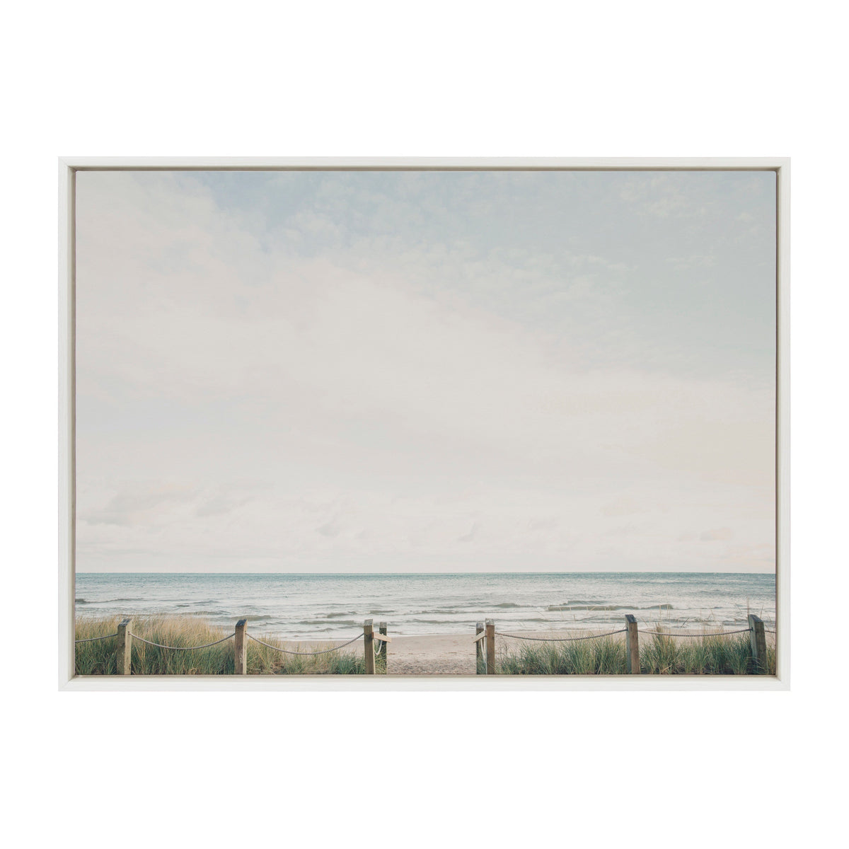 Framed canvas print showing a calm beach scene with a light blue sky, distant ocean waves, and sandy shore bordered by grassy dunes and wooden posts linked by rope.
