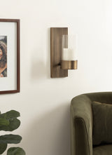 Vertical metal wall sconce with ridged brown backplate and cylindrical glass candle holder mounted above an olive green upholstered chair armrest in a light, minimalist room.