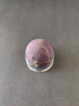 Polished purple Purpurite lapidary sphere with natural veining details, displayed upright on a clear acrylic stand against a plain gray background.
