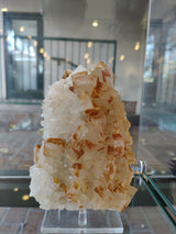 Cluster of translucent quartz crystals with scattered orange barite patches, displayed on a transparent stand against a glass case backdrop with blurred reflections.