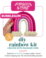 Hand holding a small rainbow-shaped macrame cord decoration in shades of pink and white with cream-colored fringe ends labeled 'Bubblegum'.