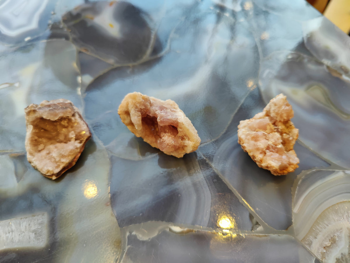 Three small pink amethyst geode clusters with crystalline formations resting on a polished stone surface featuring swirling blue, gray, and white bands.