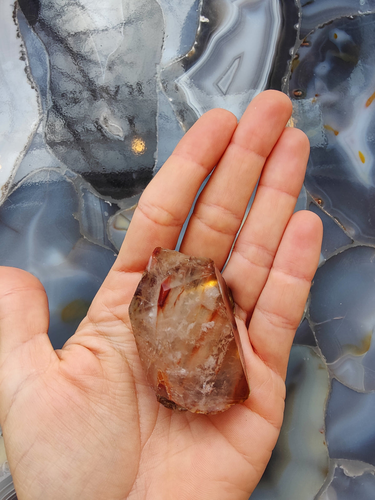 Hand holding a rough, translucent quartz crystal with reddish inclusions against a polished blue and gray agate stone background.