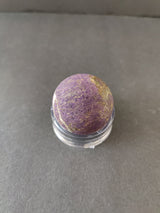 Polished purple Purpurite stone sphere with natural brownish-golden streaks, displayed on a clear round stand against a plain gray background.