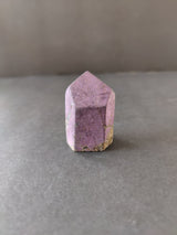 Purpurite crystal point with a faceted, hexagonal shape and a pinkish-purple hue, resting on a gray surface with natural mineral inclusions at the base.