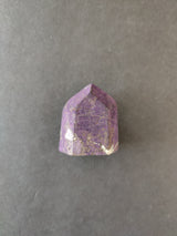 Small polished purpurite stone with purple and lavender hues and natural veining, displayed on a plain gray background.