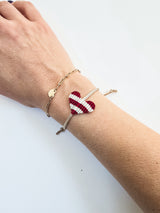 Wrist wearing an adjustable macrame bracelet with a red and white striped heart charm and a gold chain bracelet with a disc accent on a beige cord.