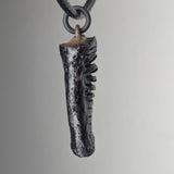 Pendant shaped like a dark textured antler or fossil with a rough surface, suspended from a black braided cord with a black ring attachment against a neutral background.