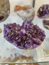 A close-up of a deep purple amethyst geode cluster featuring numerous sharp crystals with varied angular facets, displayed on a reflective glass surface with blurred stones in the background.