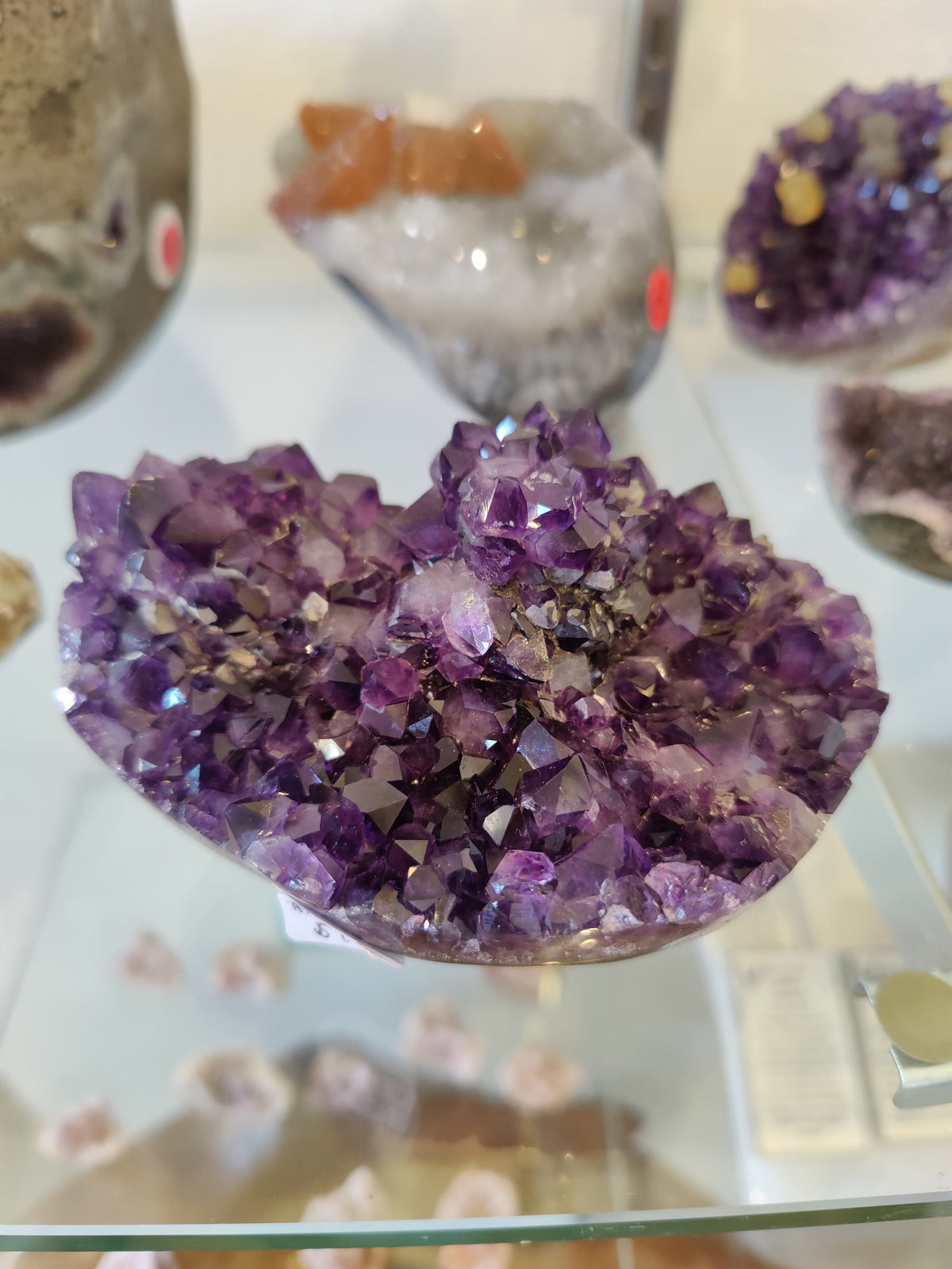 A close-up of a deep purple amethyst geode cluster featuring numerous sharp crystals with varied angular facets, displayed on a reflective glass surface with blurred stones in the background.