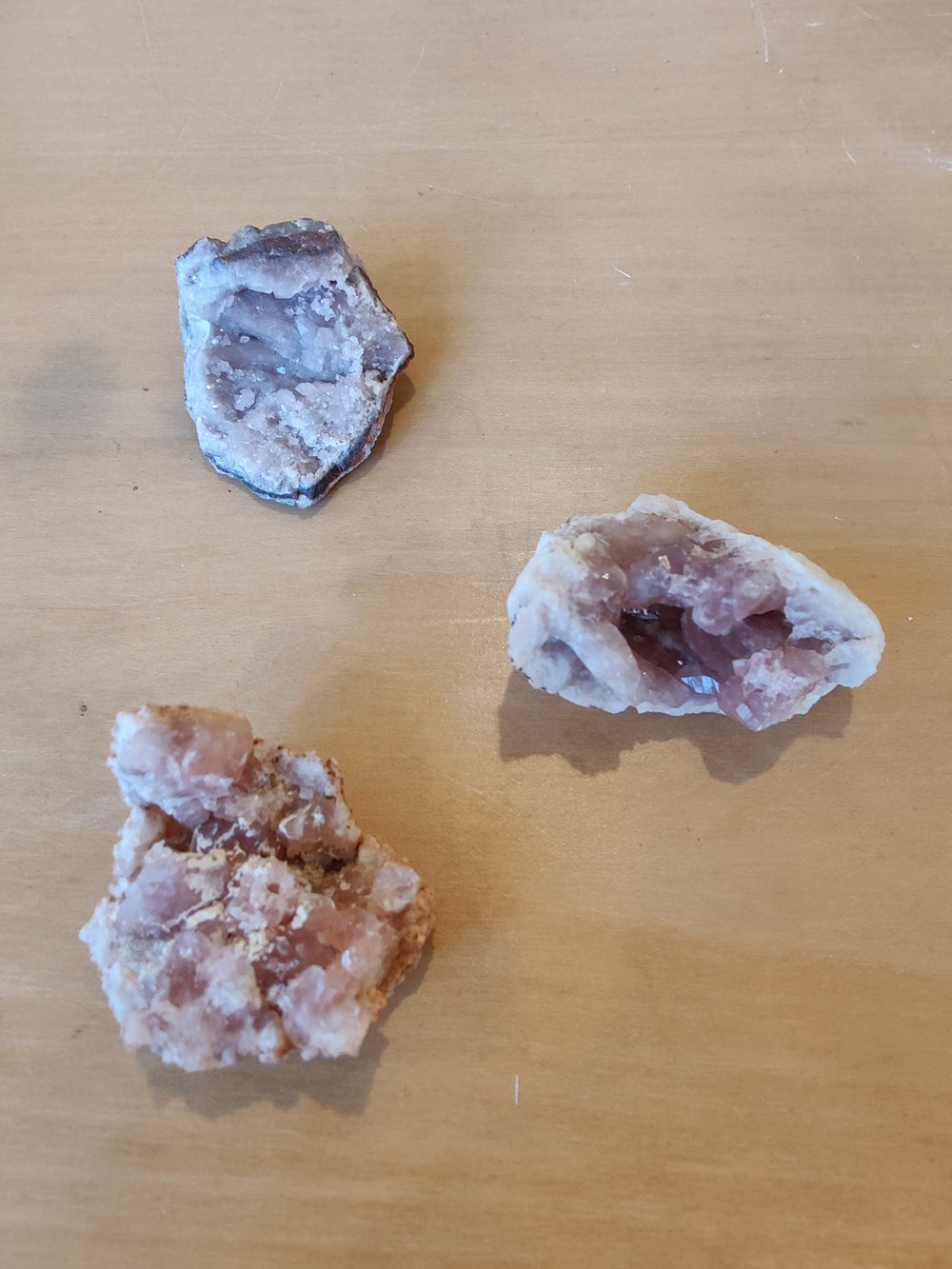 Three pink amethyst geode pieces with crystalline interiors, varying in shape and size, arranged on a smooth light wooden surface.