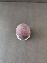 Spherical purpurite stone with purple and black marbled patterns set on a small transparent stand against a gray background.