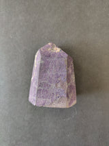 Faceted purpurite lapidary stone with a deep purple hue and visible natural matrix on a plain gray background.