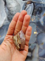 Hand holding a rough amphibole quartz crystal with brown and translucent purple hues against a polished blue and gray agate slab background.