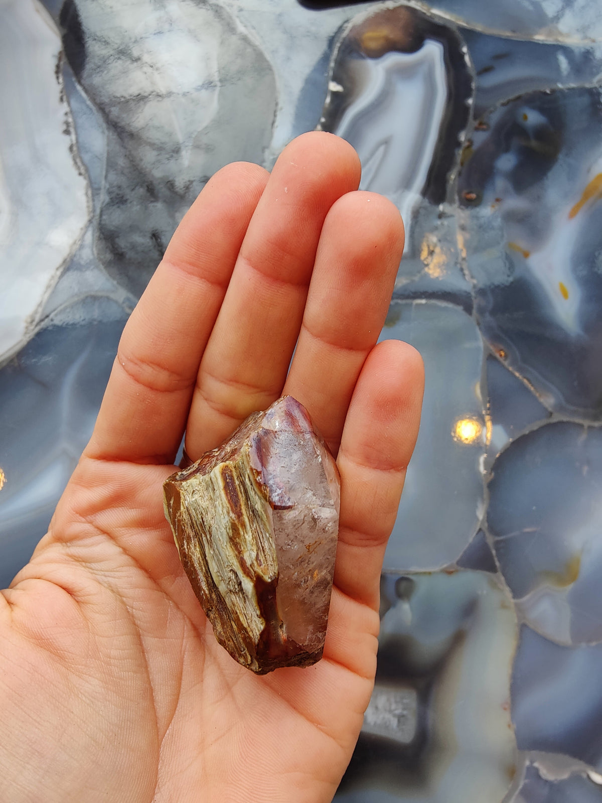 Hand holding a rough amphibole quartz crystal with brown and translucent purple hues against a polished blue and gray agate slab background.