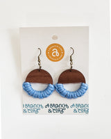Pair of earrings with dark wood semicircles topped by brass hooks and light blue macrame knotting along the semicircle edges, displayed on a white card.