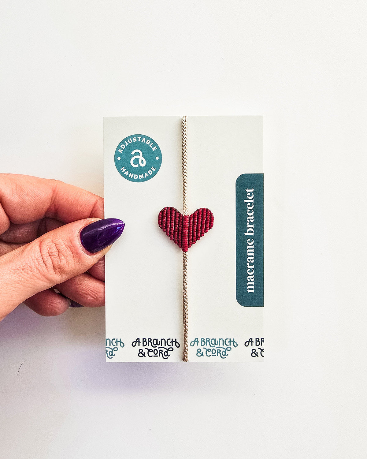 Hand holding a small card displaying an adjustable macrame bracelet with a red heart-shaped centerpiece on beige cord, labeled handmade and adjustable.