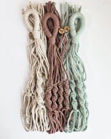 Three macrame hanging planter cords in cream, dusty rose, and sage green, each with fringed ends and wooden beads near the loop top.