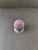 Spherical purpurite stone with purple hues and natural dark veining, displayed on a clear circular stand against a gray background.