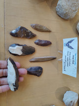 Collection of six dark brown and tan chipped stone tools of varying shapes displayed on a wooden surface, with a hand holding one at the bottom left.