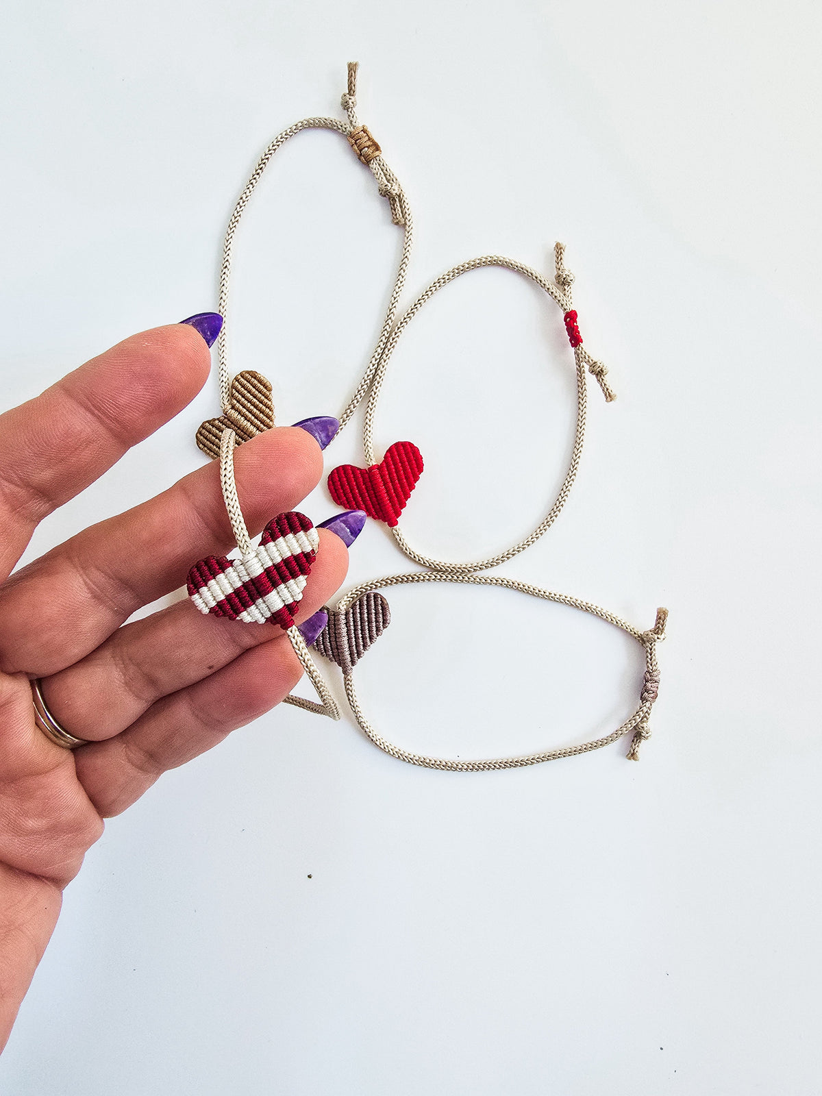 Three adjustable macrame bracelets each with a heart-shaped centerpiece in red and brown tones, held against a white background by a hand with purple nail polish.