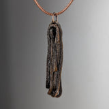 Pendant necklace featuring a dark, textured, twisted wood-like piece hanging from a thin copper-colored chain against a neutral gray background.