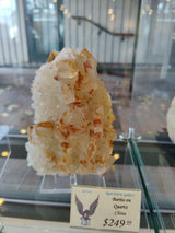 Cluster of orange-tinted barite crystals growing on a white quartz matrix displayed in a glass case with soft reflections in the background.