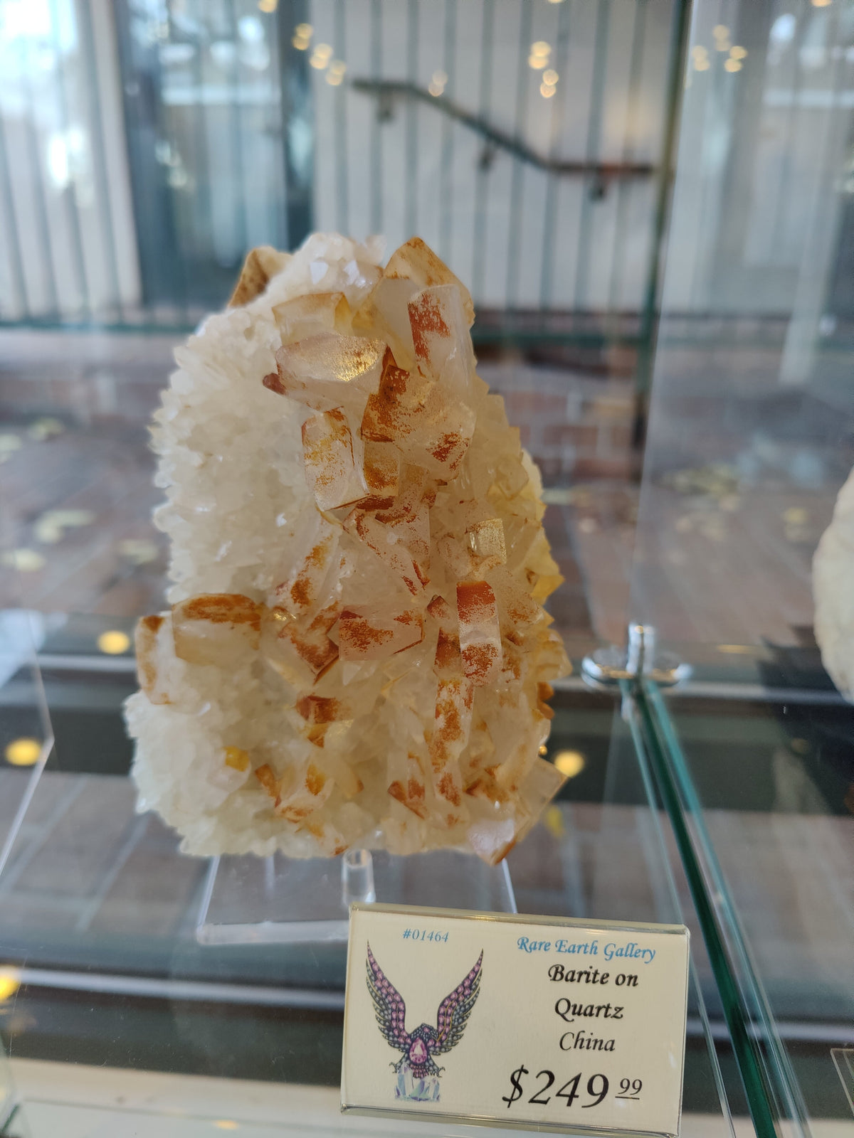Cluster of orange-tinted barite crystals growing on a white quartz matrix displayed in a glass case with soft reflections in the background.