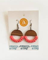 Round wooden earrings with half-circle shape featuring pink macrame knotting on the lower half, displayed on a white card with brand logo and text.