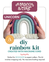 Hands holding a handmade macrame rainbow with pink, teal, and light blue arcs and white fringed ends, labeled 'Unicorn' from A Branch & Cord DIY Rainbow Kit.