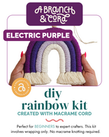 Hands holding a macrame rainbow with three arches in shades of purple and cream, each arch ending in creamy tassels.
