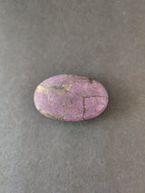 Oval-shaped purpurite stone with a smooth surface, displaying a mix of purple hues and black veining, on a plain dark gray background.