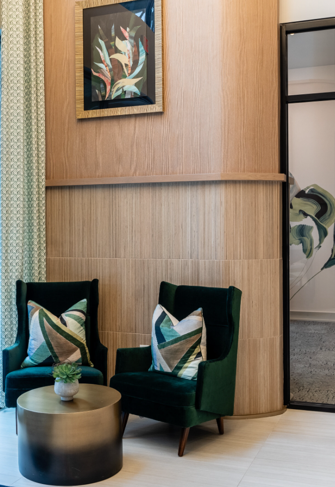 Two deep green velvet armchairs each with a geometric patterned pillow in shades of green and beige, flanking a round metallic coffee table with a small plant on top.