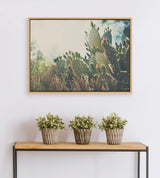 Framed canvas photograph of prickly pear cactus with green pads and red buds, hung above a minimalist wood and black metal table holding three small potted plants.