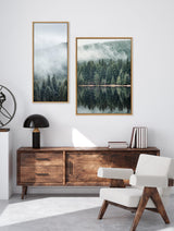 Two wooden-framed forest landscape photos with mist and green pine trees, hung asymmetrically above a mid-century modern wooden sideboard and white armchair.