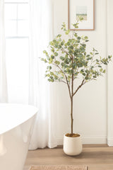 Artificial eucalyptus tree with slender brown branches and round green leaves in a white pot, standing on light wood flooring against a white wall near a window and bathtub.