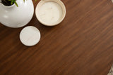 Close-up of a smooth, round wood coffee table surface in warm brown tones showing subtle grain with a white vase and two round white candles on top.