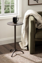 Dark metal round side table with tripod base beside olive green sofa draped with chunky cream knit blanket, natural light from window behind.