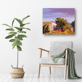 Painting of a rusty red truck near light-colored buildings and green trees under a purple and blue sky, hanging above a light gray armchair with a green throw and a potted plant beside it.