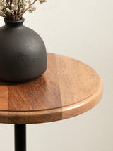 Close-up of a round wooden table top with a smooth finish, holding a matte black textured vase with dried flowers against a neutral wall background.
