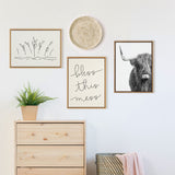 Minimalist wall gallery with three wooden-framed prints including a black line drawing of plants, 'bless this mess' script, and a black-and-white Highland cow portrait, above a light wood dresser and woven basket with pink throw.
