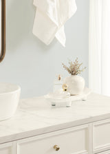 White oval decorative tray with subtle feet on white marble countertop, holding a small glass bottle with gold top and a cream vase with delicate dried flowers, under soft natural light.