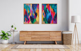 Two framed abstract canvas artworks side by side above a light wood sideboard, featuring vibrant brushstrokes in blue, pink, orange, and green hues with black and white dotted accents.