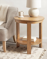 Round wooden side table with natural finish holding a white spherical lamp and mug, beside a beige upholstered armchair on a patterned rug.