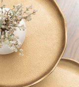 Close-up of two textured, round, gold metal trays with organic uneven edges, one partially overlapping the other on a wooden surface.
